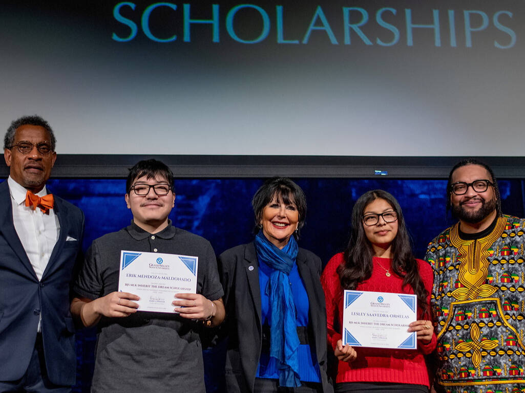 Five individuals stand on a stage under a “SCHOLARSHIPS” banner, with two holding certificates, during an awards ceremony.